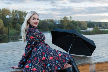 Siyah şemsiyeli bir kız, sahnedeki mavi gökyüzünün arka planında, ahşap bir merdivende oturur. Güzel bir gülümsemeyle