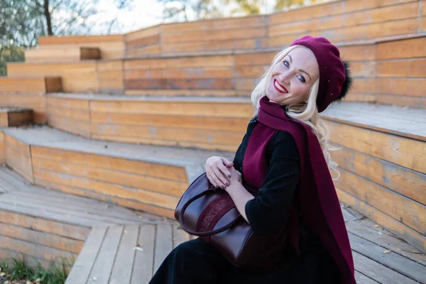 Eine fröhliche Dame sitzt auf einer sommerlichen Theaterbank aus Holz in weinrotem Mantel und Biret, eine Erwachsene, im Herbst vor blauem Himmel. — Stockfoto