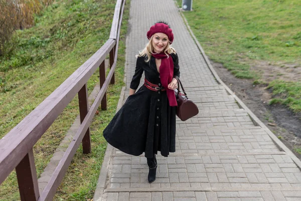 Happy walks up the stairs and in a burgundy palette and biret, an adult in black clothes, in the fall against the background of a pond a blue sky.