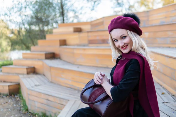 Uma mulher sorrindo senta-se em um banco de teatro de verão, feito de madeira em um casaco de Borgonha e biret, um adulto olha para a câmera, na queda contra um fundo de nuvens azuis. — Fotografia de Stock