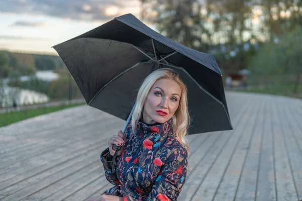Uma menina com um guarda-chuva preto senta-se em uma escada de madeira, contra o fundo das nuvens no palco. a olhar para a câmara — Fotografia de Stock