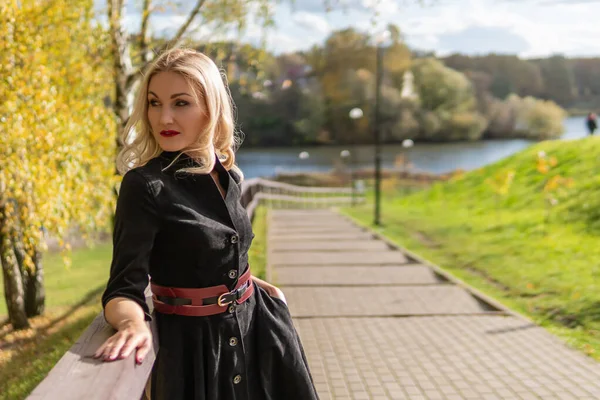 En glad dam står och lutar sig på räcket, med ett vackert leende i svarta kläder, på hösten mot bakgrund av en damm med en blå himmel. — Stockfoto