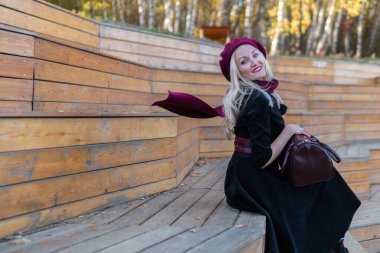 Gülen kadın yaz tiyatrosunda bir bankta oturur, bordo palto ve biet ile ahşap, sonbaharda mavi bulutların arka planında..