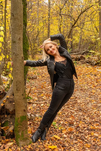En el bosque, una chica está de pie en una chaqueta de cuero y jeans negros. Hermosa sonrisa en el concepto de comparación de cámara — Foto de Stock