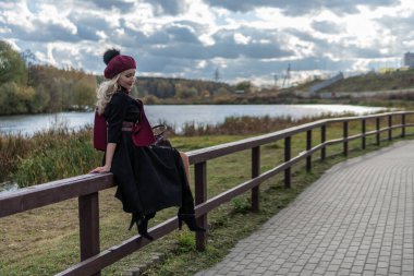 Gülümseyen bir bayan ayağa kalkar ve korkuluklara yaslanır, siyah elbiseli güzel gözleri olan bir yetişkin, sonbaharda mavi gökyüzü olan bir gölün arka planına yaslanır..