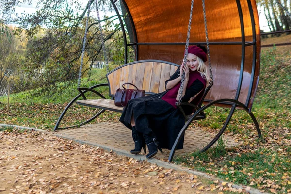 美しい目で、木、青い雲の背景に秋には、美しい少女は、バーガンディコートとビレットで鍛造、庭のスイングに座っている. — ストック写真
