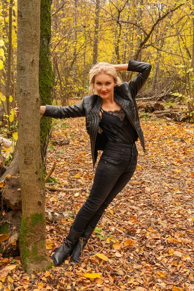 En el bosque, una chica está de pie en una chaqueta de cuero y jeans negros. Alegre mirando el concepto de cámara de la actividad webinar — Foto de Stock