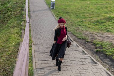 Neşeli bir kız merdivenlerden yukarı çıkar bordo palet ve biet ile, siyah elbiseli güzel bir gülümsemeyle, sonbaharda mavi bulutların arkasında.