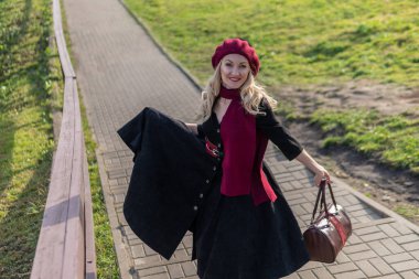 Neşeli bir kadın bordo palla ve biet ile merdivenlerden yukarı çıkar, siyah elbiseli güzel gözleriyle, gölün arka planında mavi bulutlar vardır..