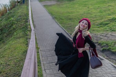 Güzel bir bayan merdivenlerden yukarı çıkıyor ve bordo bir palet ve biet ile, siyah elbiseli kameraya bakıyor, ağaçların arka planında, mavi gökyüzü.