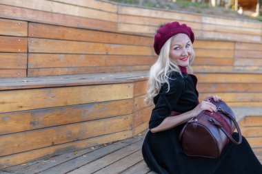 Neşeli bir yaz tiyatrosu bankında oturur, bordo bir palto ve biet içinde ahşap, kameraya gülümseyen bir yetişkin, sonbaharda bir göletin arka planında mavi gökyüzü.