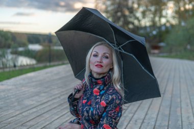 Koyu renk şemsiyeli bir kadın, sahnedeki mavi gökyüzünün arka planında, ahşap bir merdivende oturur. Nazik bir bakışla.