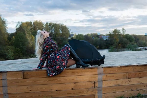 Una chica con un paraguas negro se sienta en una escalera de madera, un montón de nubes azules en el escenario. sonriendo a la cámara — Foto de Stock