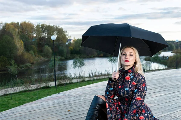 Uma mulher com um guarda-chuva preto senta-se em uma escada de madeira, contra o pano de fundo das nuvens no palco. com um sorriso adorável — Fotografia de Stock