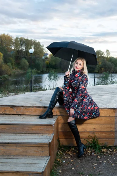 Eine Frau mit schwarzem Regenschirm sitzt auf einer Holztreppe vor dem Hintergrund eines grauen Himmels auf der Bühne. mit einem schönen Lächeln — Stockfoto