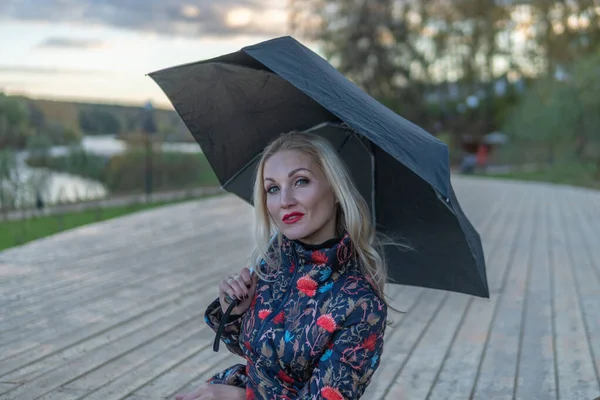 Uma menina com um guarda-chuva escuro senta-se em uma escada de madeira, contra o fundo das nuvens no palco. a olhar para a câmara — Fotografia de Stock