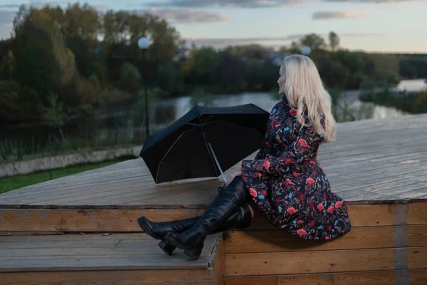 Dziewczyna z czarnym parasolem siedzi na drewnianych schodach przeciwko szaremu niebu na scenie. patrząc w kamerę — Zdjęcie stockowe
