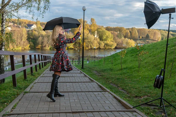傘を差した大人の女の子が秋のジャケットで堤防のフェンスに沿って立っています。黄色の森、緑の草、ブロンド。カメラの健康の概念を見ると — ストック写真