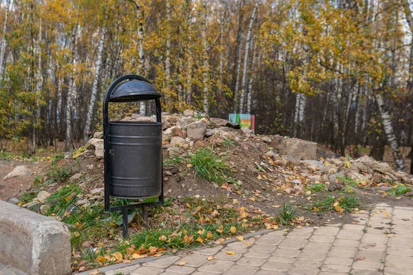 Lixeira no parque, uma lata de lixo feita de metal em um gramado em uma natureza aberta perto de uma estrada de pedras, a tarde em tempo ensolarado — Fotografia de Stock