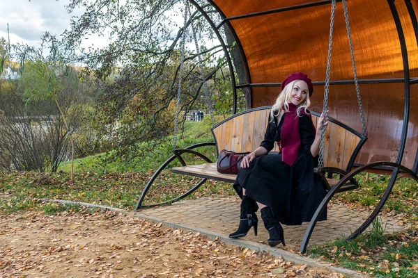 Uma mulher sorrindo senta-se em um balanço do jardim, forjado em um casaco de Borgonha e biret, com belos olhos, na queda contra o fundo das árvores, nuvens azuis. — Fotografia de Stock