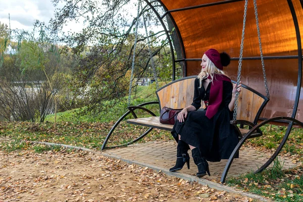 Dívka sedí na zahradním houpačce, ukována ve vínově zbarveném kabátě a biretu, usmívá se do kamery, na podzim na pozadí rybníka, modré oblohy. — Stock fotografie