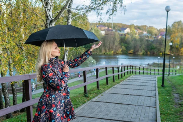 Uśmiechnięta młoda dziewczyna z parasolem stoi na płocie na nabrzeżu w kurtce jesienią. Żółty las, zielona trawa, blondynka. patrząc na koncepcję dobrego samopoczucia kamery — Zdjęcie stockowe