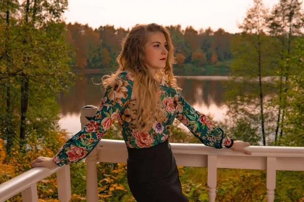 a teenager stands leaning on a white railing an adult with beautiful eyes blonde in a colored shirt positive, emotions on a lake background autumn haircut