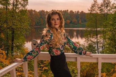 Neşeli kadın, beyaz korkuluklara yaslanmış, güzel gözlü, renkli gömlekli, sarışın, gölün arka planında, gülen sonbahar şehvetli maske maskesine yaslanmış, koyu mavi gözlü bir yetişkin.