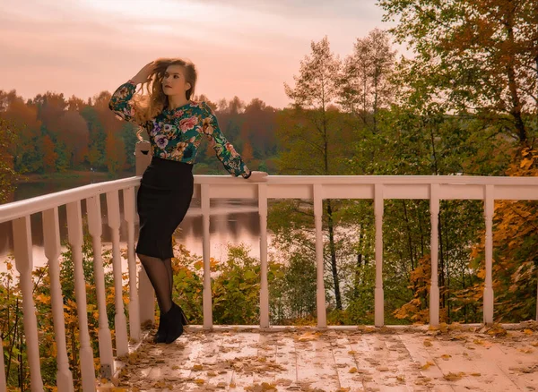 Uma bela mulher está apoiada contra um corrimão branco uma jovem loira com lábios suaves, uma camisa colorida com olhos azuis no fundo do lago fazer uma tendência sensual outono — Fotografia de Stock