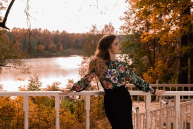 Neşeli kız beyaz bir korkuluğa yaslanmış genç kameraya bakıyor. Renkli gömlekli sarışın, sonbahar merdivenlerinin arka planında duruyor. Parlak mavi gözler.