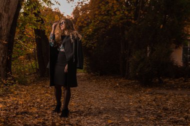 Siyah pelerinli neşeli bir kız sonbaharda bukalemun gözlüğü takıyor. Sarı orman, yeşil çimen, sarışın. Kamera kentsel yaşam konseptine bakıyorum.