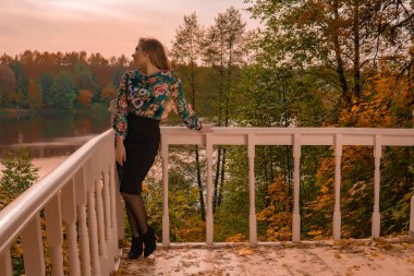 Renkli bir gömlekle karizmatik görünümlü neşeli genç bir kadın beyaz korkuluklara dayanan beyaz korkuluklara karşı duruyor. Sonbaharı şehvetli yapmak için gölün arka planında duruyor.