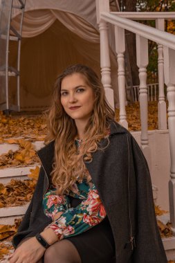 Gülümseyen bir kadın, siyah gömlekli yetişkin bir sarışının beyaz korkuluğuna yaslanmış duruyor. Adımların arka planına bakıyor. Sonbahar parlak mavi gözleri var.