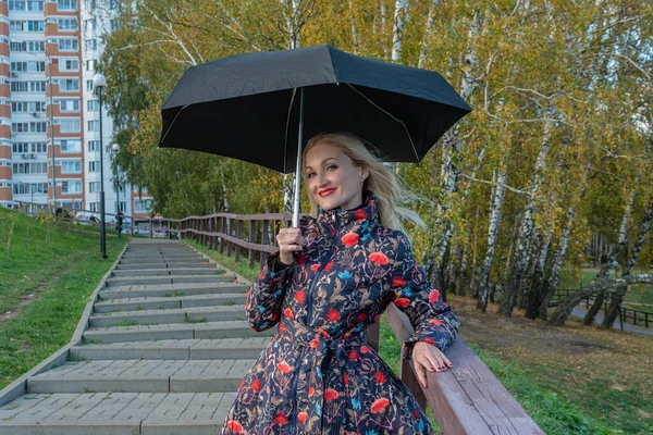 Usmívající se mladá dívka s deštníkem stojí na schodišti v kabátě na podzim. Žlutý les, zelená tráva, blondýna. s dobrým vzhledem koncept pohody — Stock fotografie
