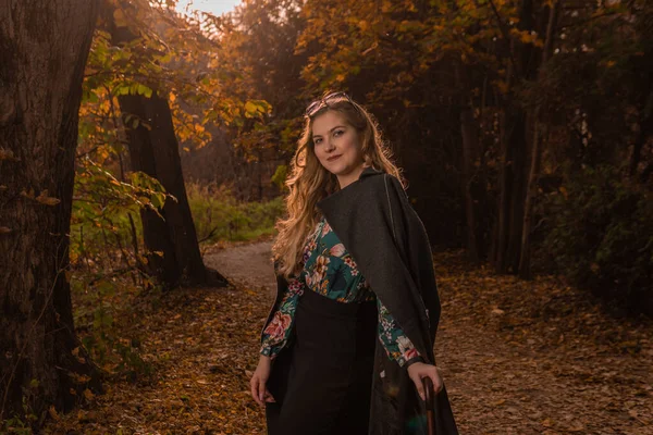 黒いマントを着た美しい少女が、秋にガラスのカメレオンの中に立っている。黄色の森、緑の草、ブロンド。カメラの都会の生活のコンセプトで — ストック写真