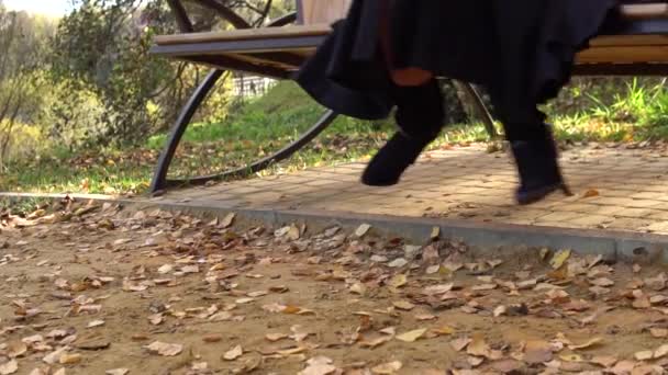 Una mujer se sienta en un columpio de jardín, arrojado en un abrigo de color burdeos y en un biret, un adulto con hermosos ojos, en otoño contra las nubes azules del estanque. Balanceo de pies visibles — Vídeos de Stock