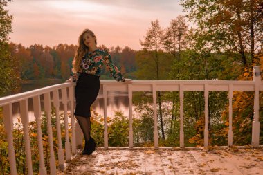 Mutlu kadın beyaz bir korkuluğa yaslanmış duruyor. Renkli bir sarışına bakıyor. Arka planda, masum bir sonbahar kolejinin parlak mavi gözlerine bakıyor.