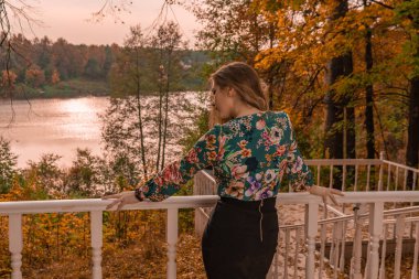 Gülümseyen genç, beyaz bir korkuluğa yaslanmış siyah tişörtlü, kamera sarışına bakıyor. Masumane sonbahar koleji maskesinin arka planında duruyor.