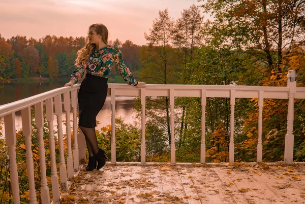 Adolescente sonriente de pie apoyado en una barandilla blanca mirando a la cámara rubia, en una camisa de color que tiene salón de otoño inocente en el fondo del lago de pie máscara oscura ojos azules — Foto de Stock