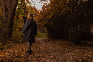 Sonbaharda siyah pelerinli genç bir kız bukalemun gözlüğü takıyor. Sarı orman, yeşil çimen, sarışın. Karizmatik görünümlü. Konseptin tadını çıkar.