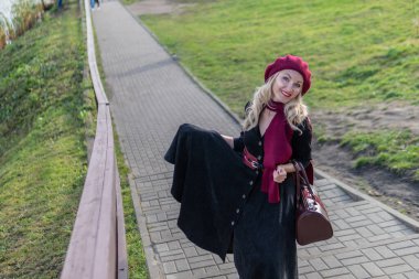 Gülümseyen bir bayan merdivenlerden yukarı çıkıyor ve bordo palet ve biet giyiyor, siyah elbiseli kameraya gülümsüyor, sonbaharda mavi bulutlu bir gölün arka planında..