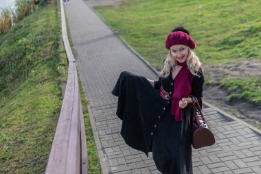 Gülümseyen bir kadın merdivenlerden yukarı çıkıyor ve bordo bir palet ve biet giyiyor, sonbaharda mavi gökyüzüne bakan siyah elbiseli kameraya bakıyor..