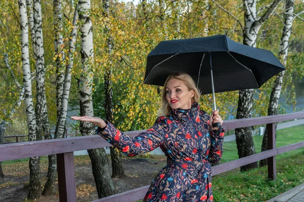Uma jovem alegre com um guarda-chuva fica ao longo da cerca no aterro em uma jaqueta no outono. Floresta amarela, grama verde, loira. olhando para o conceito de comparação de câmera — Fotografia de Stock
