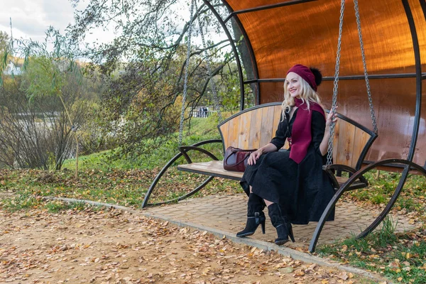 Veselá dáma sedí na zahradním houpačce, ukována ve vínovém kabátě a biretu, s krásnýma očima, na podzim na pozadí rybníka je modrá obloha. — Stock fotografie