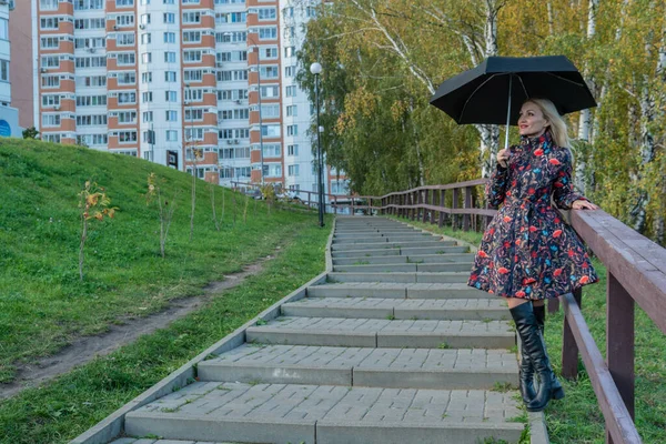 傘を差した陽気な少女が、秋のジャケット姿で階段の上の人形の柵の中に立っている。黄色の森、緑の草、ブロンド。美しい目で都市生活の概念 — ストック写真