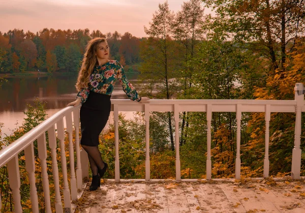 Una chica alegre se apoya sobre una barandilla blanca un joven con una mirada amable rubia, en una camisa de color mira el fondo del lago risa otoño tendencia sensual — Foto de Stock