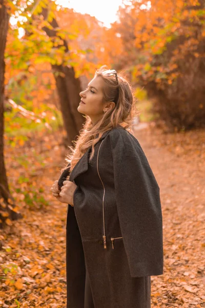 Una hermosa joven con una capa negra está de pie en los camaleones de gafas en el otoño. Bosque amarillo, hierba verde, rubia. con concepto de comparación de labios tiernos — Foto de Stock