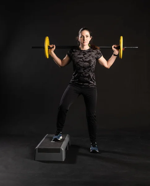 Fitness en una plataforma escalonada, con una barra, tortitas amarillas tirando de una pierna. se dedica a los deportes sobre un fondo negro hermosa mirando a la cámara — Foto de Stock