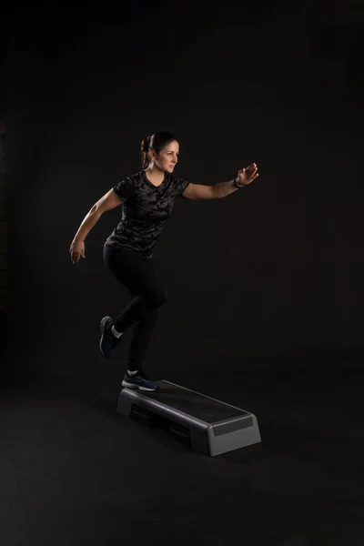 Chica de fitness en una plataforma escalonada, tira de su pierna. Ir en los deportes sobre un fondo negro Alegre — Foto de Stock