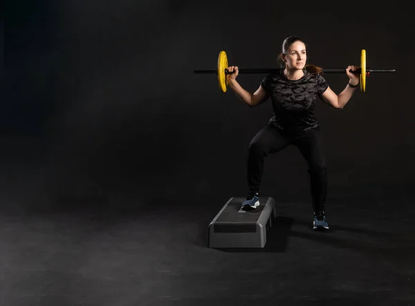 Fitness em uma plataforma de passo, com um sino de barra, panquecas amarelas mantém o equipamento. Está envolvido em esportes em um fundo preto Bonito sorrindo para a câmera — Fotografia de Stock